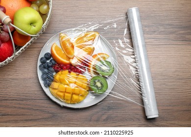 Plate Of Fresh Fruits And Berries With Plastic Food Wrap On Wooden Table, Flat Lay