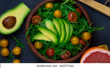 Plate With Fresh Salad With Arugula, Cherry Tomatoes And Avacado, Bright Green On A Black Background, Avacado And Grapefruit Cut In Half, Top View