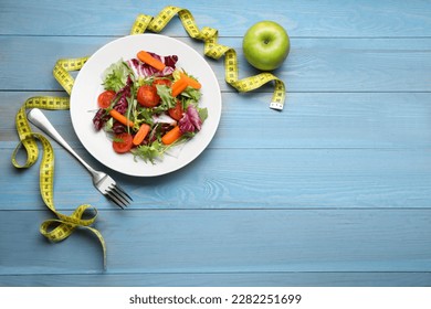 Plate of fresh vegetable salad, apple and measuring tape on light blue wooden table, flat lay with space for text. Healthy diet concept
