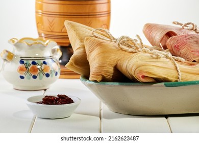 Plate full of salty and sweet tamales wrapped in corn leaves by a typical Mexican chocolate cup and some chipotle chili peppers over a white wooden table.