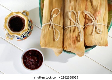 Plate full of tamales wrapped in corn leaves by a typical Mexican chocolate cup and some chipotle chili peppers over a white wooden table. Top view.