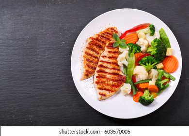 plate of grilled chicken with vegetables on dark background, top view
