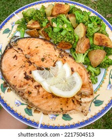 A Plate Of Grilled Salmon Steak With Some Lemons Slices And Pan Fried Broccoli And Roasted Potatoes.