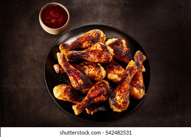 Plate of grilled spicy chicken legs served ready for a finger buffet with a side dish of hot chili sauce, overhead view on a slate background