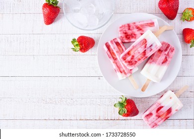 Plate of homemade strawberry vanilla yogurt popsicles. Above view corner border on a white wood background with copy space.