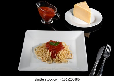 A Plate Of Pasta, With Red Sauce,
 Next To The Plate A Container With Sauce And A Piece Of Parmesan Cheese On A Dark Glass Table