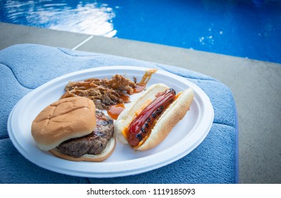 A plate of pulled pork, cheeseburger, and a hot-dog are displayed poolside. 