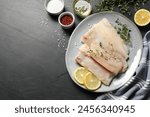 Plate with raw cod fish, microgreens, spices and lemon on dark textured table, flat lay. Space for text