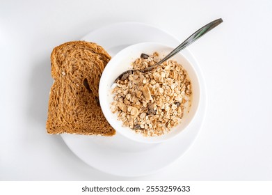 Plate with simple breakfast centered in the frame. Bowl with muesli and yogurt and a slice of brown bread next to it. White soft background with natural light.