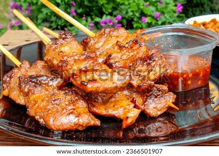 a plate of skewers of meat and a bowl of sauce on a wood table, with a garden setting in the background