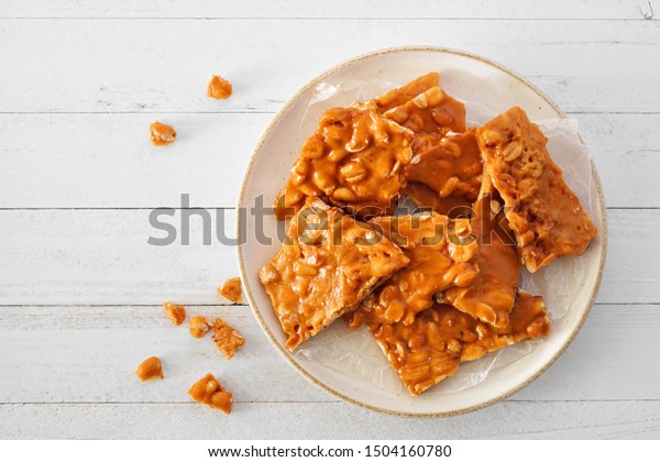 Plate of traditional peanut brittle\
candy pieces. Top view on a white wood\
background.