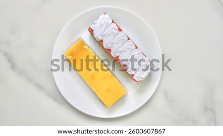 A plate with two rectangular desserts—one is a cheesecake with a glossy yellow topping, and the other is a pink pastry with piped white cream.                    