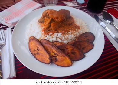 A Plate Of White Rice And Fried Plantain Served With Nigerian Red Spicy Stew, Chicken And Kpomo Meat. Beautifully Set On A Restaurant Table.