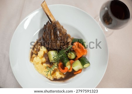 A plated fine dining dish featuring a herb-crusted lamb chop with bone, served with creamed potato, saut?ed courgette, red pepper, and fresh rosemary garnish, accompanied by red wine.