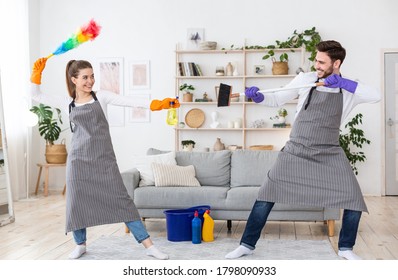 Play And Fighting During Cleaning. Married Couple In Aprons And Rubber Gloves Have Fun In Living Room Interior With Cleaning Supplies, Free Space