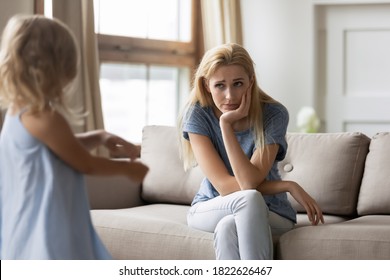 Play with me! Weary young woman babysitter hiding on couch with tired unhappy look from naughty active girl demanding to play, distressed mom with a pity face listening to capricious little daughter