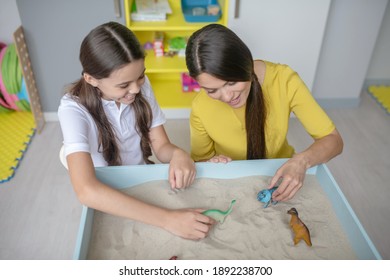 Play, Therapy. Smiling Cute Dark-haired Woman And Girl Rearranging Small Figures On Sand In Tray In Playroom