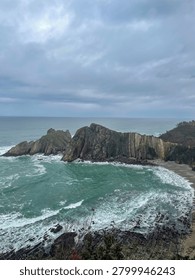 Playa de Silencio dramatic vertical rock strata rising from turquoise waves, with a secluded sandy beach, rugged jagged rocks and a moody overcast sky, Cantabrian coast, Asturias, Spain.