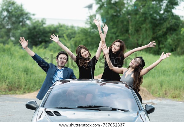 Playboy party concept. Young white male\
inside convertible sport car with four beautiful asian women doing\
a freedom pose while going to a carefree\
party.
