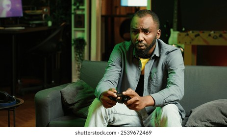 Player in RGB lit home gaming with joystick and enjoying relaxation time. Man sitting on sofa in neon illuminated living room, playing videogames on console using controller, camera A - Powered by Shutterstock