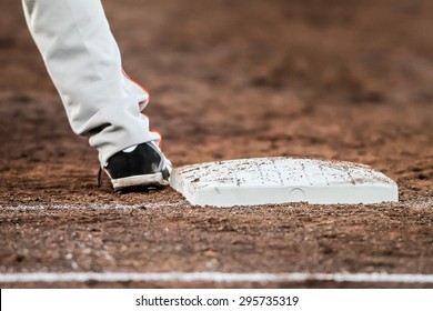 Player Waiting With Hes Feet Touching The Base Plate Infield During A Baseball Match. The Baseball Player Wears Black Shoes Touching The White Baseplate Laying In Brown Dirt.