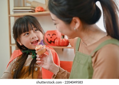 Playful And Adorable Young Asian Girl In A Halloween Fancy Costume Is Eating A Cupcake With Her Mom In The Kitchen. Close-up Image