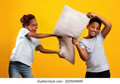 Playful African American Teen Sister And Brother Having Pillow Fight Over Yellow Studio Background, Happy Black Siblings Or Twins Having Fun, Enjoying Time Together, Isolated. Siblings Relationships