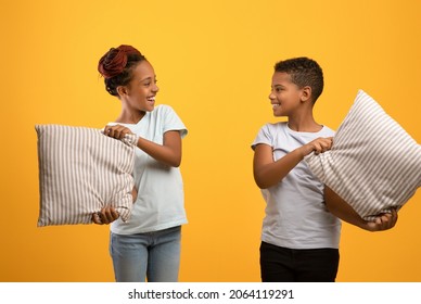 Playful African American Teen Sister And Brother Holding Pillows Over Yellow Studio Background, Cheerful Black Siblings Or Twins Having Pillow Fight, Enjoying Time Together. Siblings Relationships