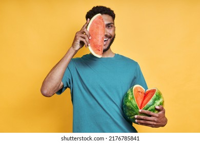 Playful African man holding slice of watermelon near face while standing against yellow background