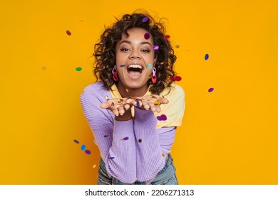 Playful African woman blowing confetti and smiling while standing against yellow background - Powered by Shutterstock