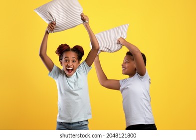 Playful Afro-american Teen Sister And Brother Holding Pillows Over Yellow Studio Background, Cheerful Black Siblings Or Twins Having Pillow Fight, Enjoying Time Together. Siblings Relationships