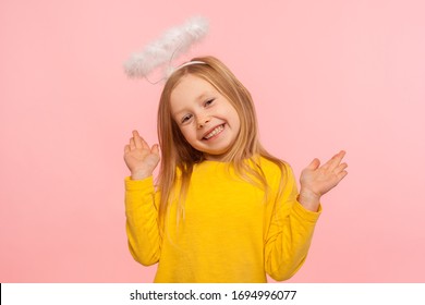 Playful Angelic Little Girl Raising Arms And Looking At Camera With Toothy Smile, Gesturing Oops, I Don't Know, It Wasn't Me, Joyful Child Denying Guilt. Indoor Studio Shot Isolated On Pink Background