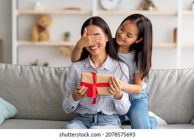 Playful Asian Girl Making Surprise For Her Happy Mother, Closing Her Eyes And Giving Gift Box While Sitting On Couch At Home Interior, Copy Space