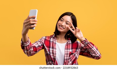 Playful Asian Lady Taking Selfie On Smartphone And Showing Peace Gesture At Webcamera, Korean Woman Having Fun While Making Photo Over Yellow Background, Panorama