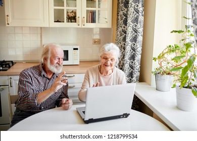 Playful Beautiful Mature Couple Of Pensioners Sitting At Kitchen Table And Watching Video From Their Wedding Anniversary Using Laptop.