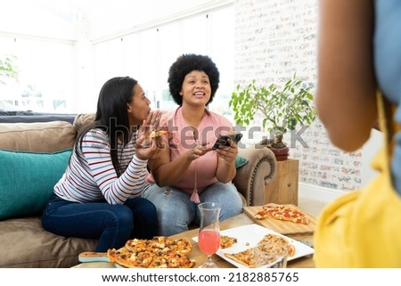 Playful biracial woman eating pizza and teasing friend using cellphone while sitting on sofa at home. Technology, food, unaltered, togetherness, social gathering, enjoyment and weekend activities.