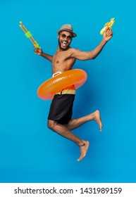 Playful black man jumping with water guns, enjoying summer time and pool party, blue studio background