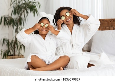 Playful Black Mother And Little Daughter In Bathrobes Applying Cucumber Pieces To Eyes, Having Fun Together After Bathroom, Cute Girl And Mom Sitting On Bed, Enjoying Home Beauty Spa Day, Copy Space