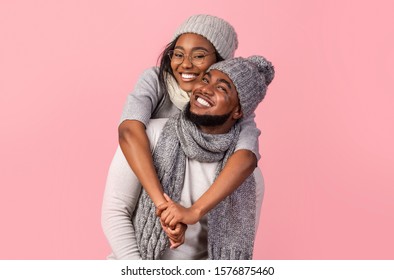 Playful black winter girl hugging her cheerful boyfriend from behind, pink background - Powered by Shutterstock