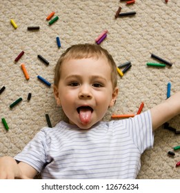 A playful boy sticks his tongue out as he lies on the floor, with crayons strewn all over.