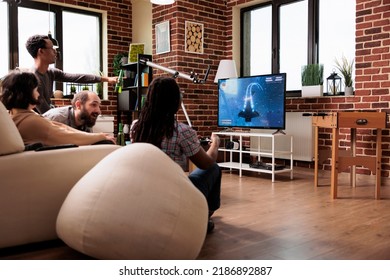 Playful Casual Multiracial People Sitting Playing Together Space Simulator Videogame On Gaming Console. Diverse Friends In Living Room On Sofa Relaxing With Games Played On Entertainment Device.