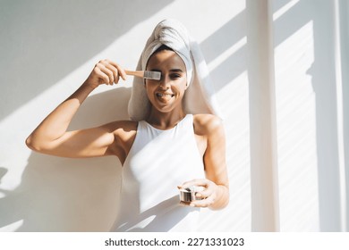 Playful Caucasian lady in white bodysuit and turban applying cosmetic cream with brush and showing tongue while standing near sunlit wall after morning shower