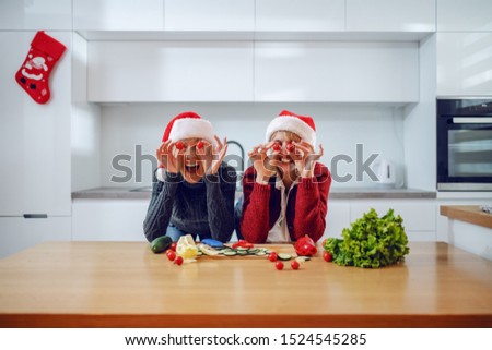 Playful caucasian woman and her mother holding tomatoes in front of their eyes while leaning on kitchen counter. Both having santa hats on heads. New year's eve.