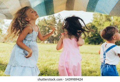 Playful Children Playing Under The Blanket, Jumping And Dancing Together. Happy Little Boy And Little Girls Enjoying Summer Day In The Park. Cheerful Kids Playing Outdoors. Childhood Concept