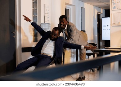 Playful corporate employee riding desk chair having fun during moving activity in startup office. Cheerful managers taking break from work enjoying doing sport races late at night. Business concept