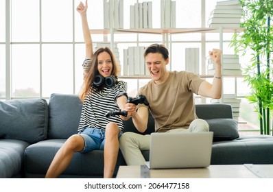 Playful Couple Hold Gamepad Controller Playing Video Games As Home Entertainment. Happy Excited Two Friends Sit On Couch In Living Room Having Fun With Videogame Hobby Leisure Indoor Activity Together