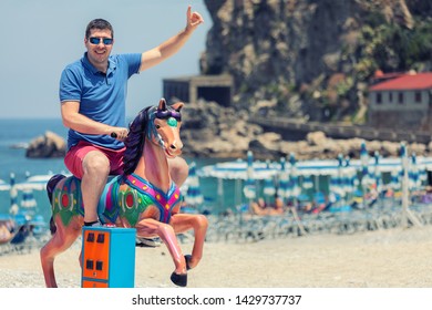 Playful Crazy Man (dad) Riding Wooden Rocking Horse At Beach – Happy Adult Guy Having Fun On Playground In Summer Vacation Playing Like Children