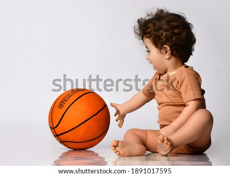 Playful curly little boy, dressed in a bodysuit, sits barefoot and plays new basketball on a gray background. Place for text. The concept of an active childhood.