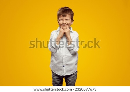 Playful cute kid wearing white shirt, touching cheeks with both index finger against yellow background, making funny grimace