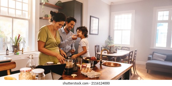 Playful dad feeding his son a slice of bread while his wife prepares breakfast. Family of three having fun together in the kitchen. Mom and dad spending quality time with their son.